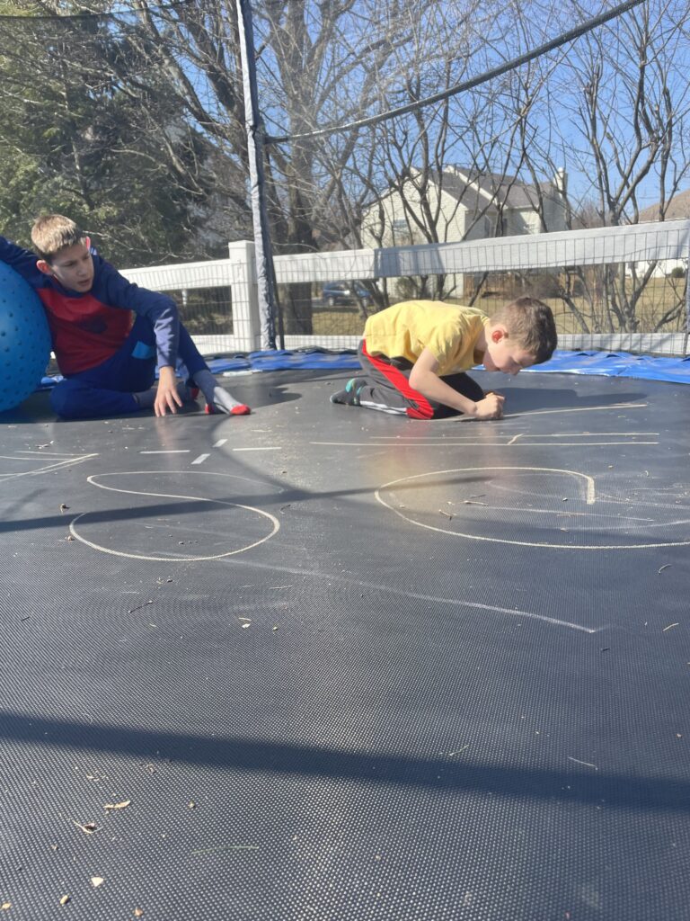 Two boys on a trampoline writing spelling words, using movement and novelty to support task initiation during homeschool.